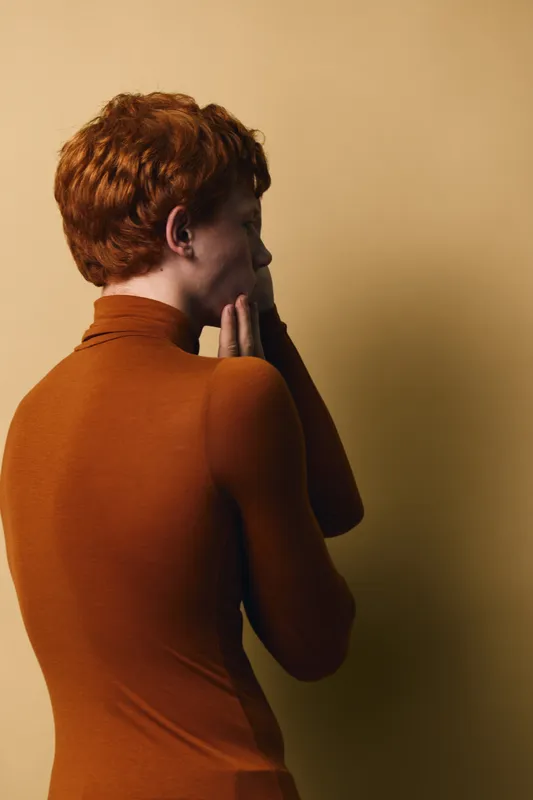 Woman with short red hair in brown turtleneck stands in profile with hand on face against beige background. Portrait, contemplation and indoor lighting for mood and style concept.