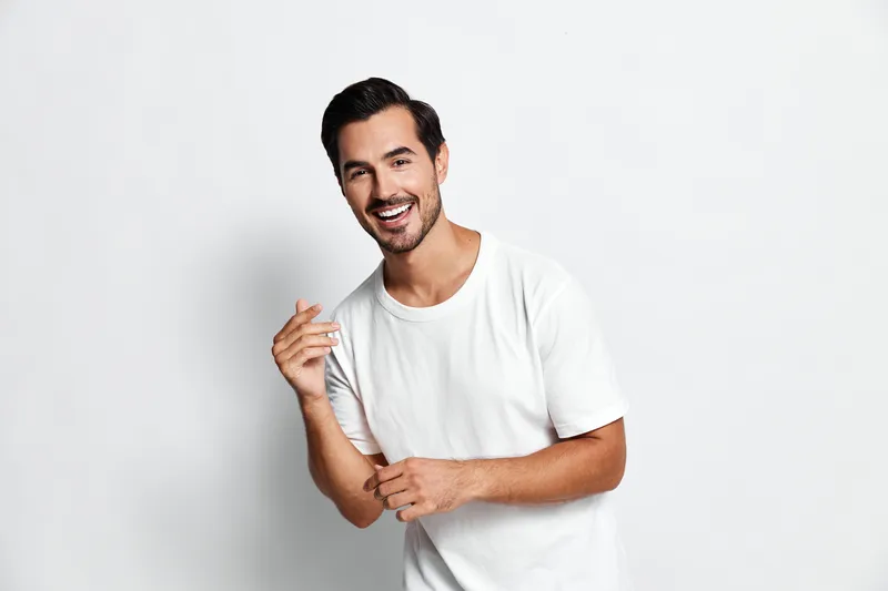 Smiling happy cheerful young man in casual white t-shirt looking at the camera with a joyful expression, standing against a plain light background.