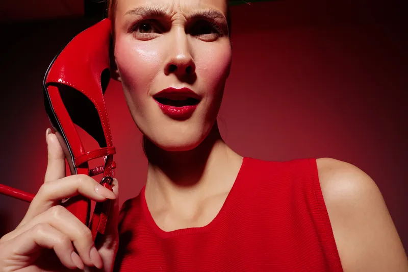 Close-up of a woman in red dress holding a red high heel shoe near her face with a curious expression. Red background enhances the bold and stylish concept of fashion and beauty.
