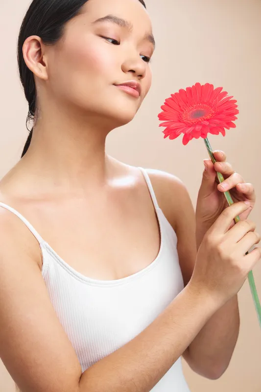 asian woman in a white tank top holds a bright flower, gazing softly at it, capturing a serene moment of beauty and calm, conveying grace and gentle self care in a simple setting.