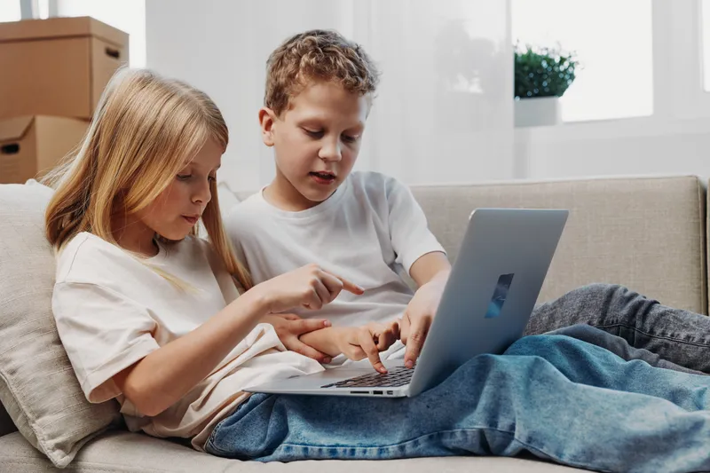 Two children sitting on a sofa using a laptop together in a cozy living room. The boy and girl focus on the screen, sharing an educational or entertainment moment at home.