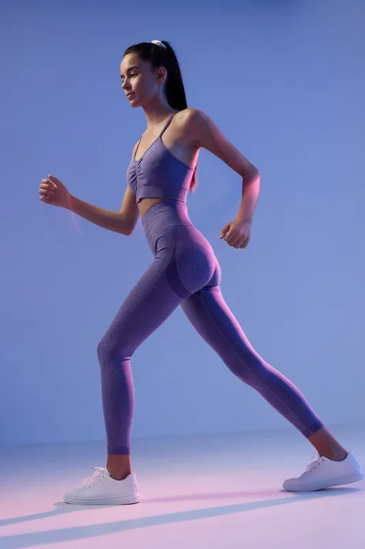 Young woman sport fitness training in purple workout clothes and white sneakers, exercising indoors with focused expression against a gradient blue background.