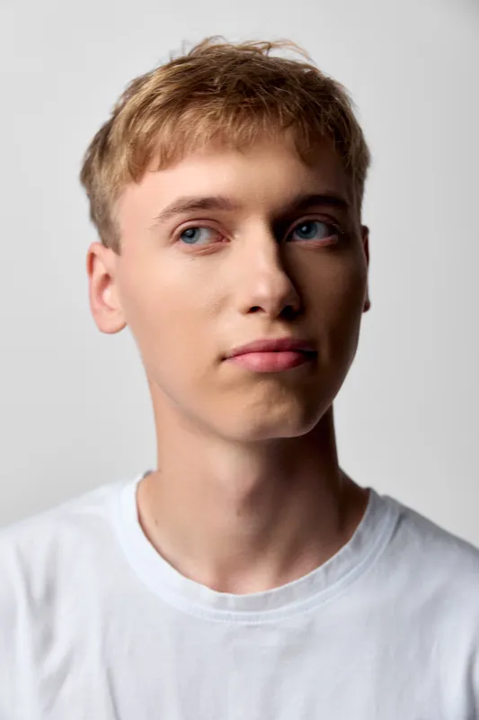 Young man with light skin wearing a casual white shirt in a studio, looking slightly to the side with a calm expression and clean, minimal backdrop