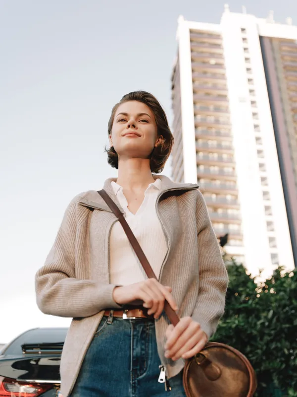 A stylish young woman walks confidently in an urban setting, looking ahead with a calm expression. She wears a light jacket and jeans, carrying a round bag, embodying independence and modern city