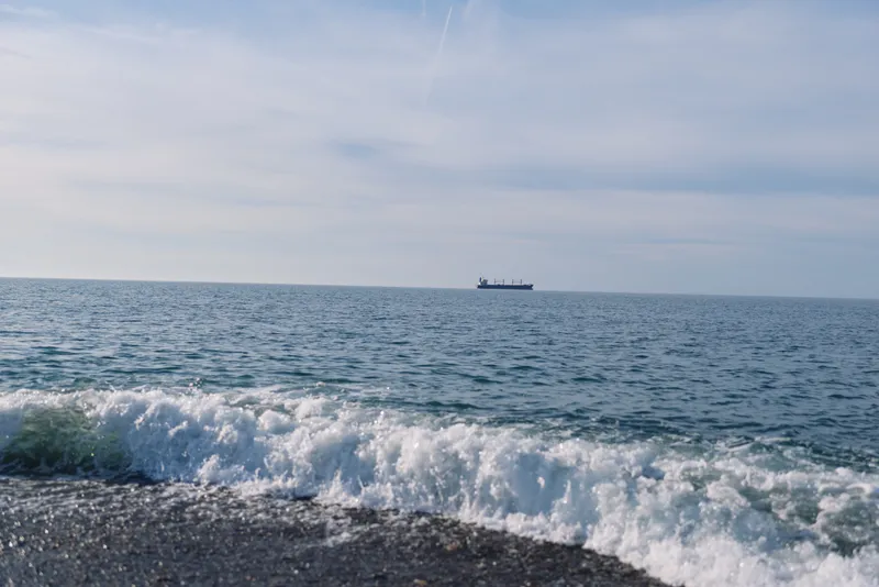 Calm ocean scene with rolling waves crashing on a pebbled shore as a distant ship glides toward the horizon under a soft, pale sky, the surface glittering and a gentle maritime mood across the
