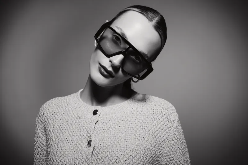 Stylish Caucasian woman with pale skin wearing large sunglasses and textured sweater poses against plain background. Fashion portrait concept.