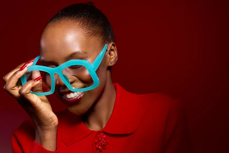 Smiling dark skinned African woman with short hair wearing large turquoise glasses and bright red clothing on red background. Concept of fashion, style, beauty, and confidence.