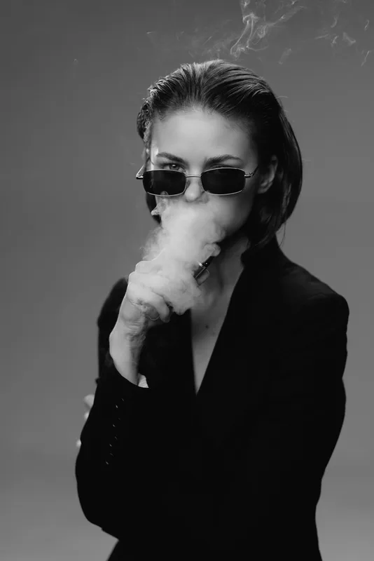Black and white portrait of a stylish woman wearing sunglasses exhaling smoke in a dark jacket, mysterious and cool atmosphere, monochrome fashion concept.