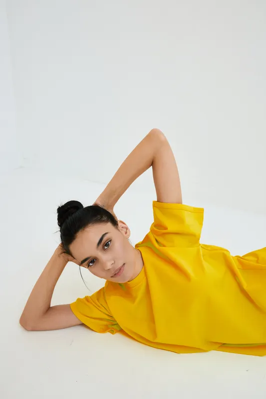 Young woman lying down in bright yellow shirt against white background, relaxed pose with arms behind head and serene expression, casual minimalistic style.