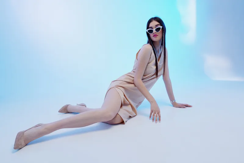 Fashionable woman in a beige dress and sunglasses poses on a light blue background. She sits elegantly with one leg extended, showcasing beauty, style, and modern fashion concepts.