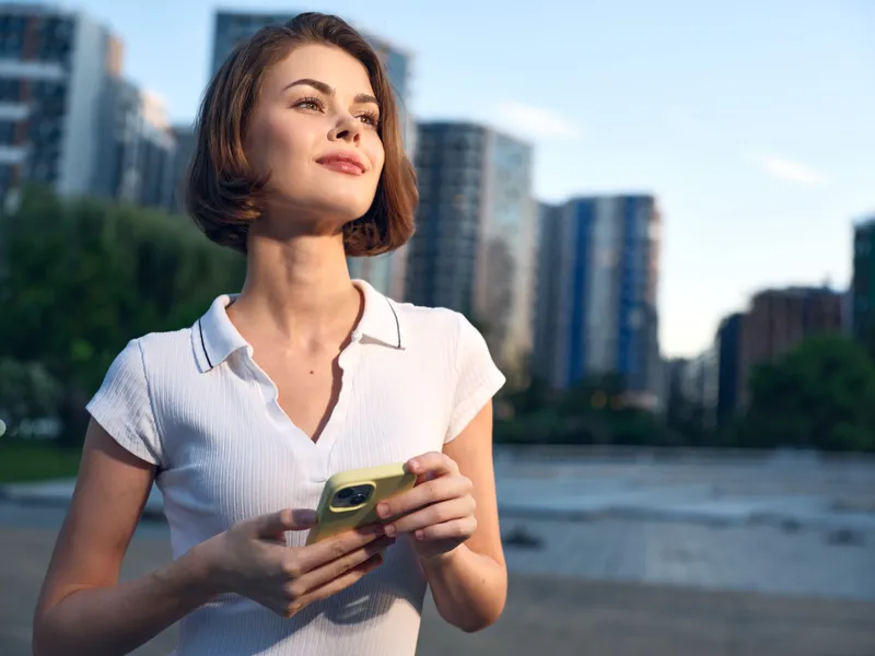 A cheerful young woman stands outdoors in an urban park, texting on a smartphone. She wears a white polo, smiles confidently, and enjoys a bright, relaxed moment.