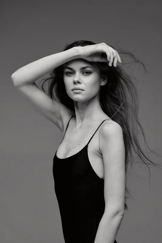 Black and white portrait of a young woman with long hair posing confidently in a sleeveless top against a simple dark background.