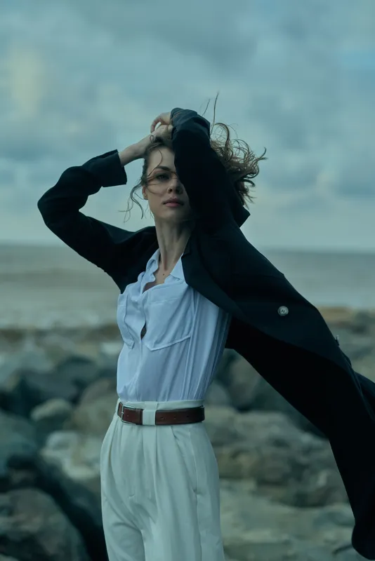 A windswept woman on a rocky beach adjusts her dark coat over a white blouse and belt, creating a dramatic fashion moment by the sea, as waves crash nearby