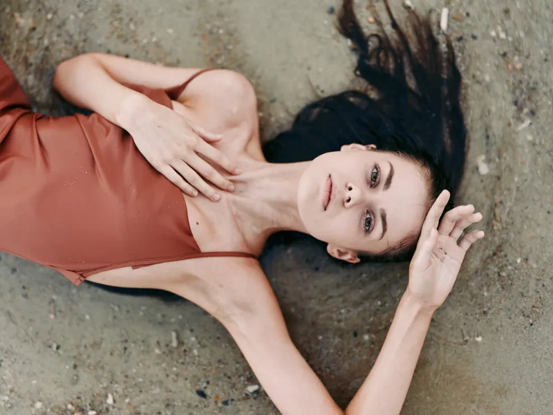 Portrait of a relaxed woman lying on sandy beach in a rust swimsuit, sunlit skin and soft shadows create a serene, intimate mood and natural casual pose.