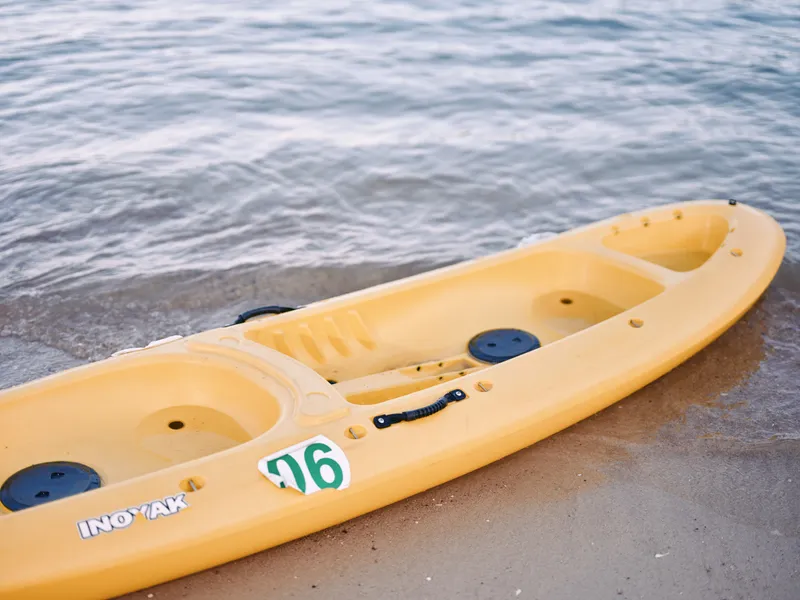 Bright yellow kayak rests on the sandy shore beside calm water, featuring molded seats and grab handles, inviting outdoor exploration, leisure paddling, and sunny seaside moments.