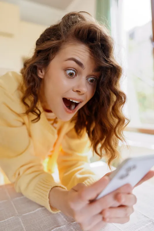 Candid woman with wavy hair wearing a yellow sweater, looking surprised while scrolling her phone. Cozy home setting with soft light, capturing playful daily life moment.