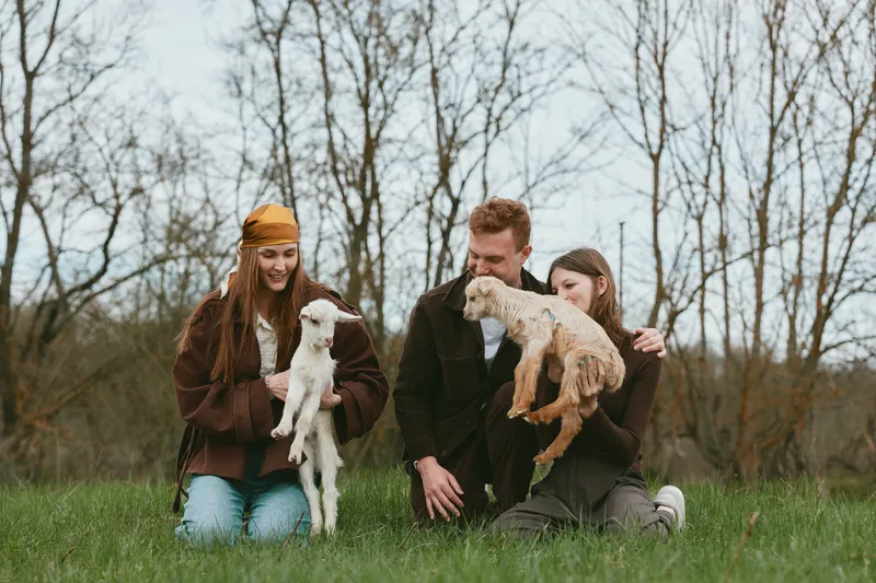 Cheerful family outdoors with goats, sharing a candid moment on a grassy field, casual jackets, natural light, warm breeze, relaxed posture, rustic backdrop, affectionate interaction, wholesome