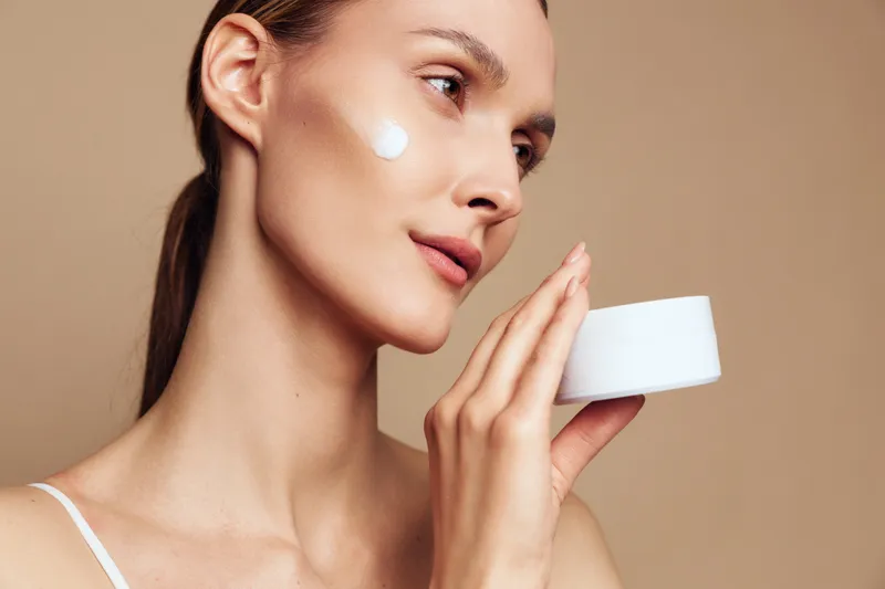 Woman skincare face beauty cream moisturizing spa studio, young woman applying moisturizer to cheek, soft light, calm expression, relaxing self care, indoor beige background, close up