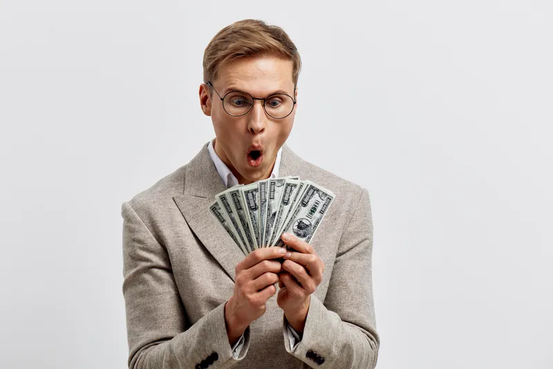 Confident man in glasses holding a large fan of dollar bills with surprised expression, dressed in formal beige blazer, isolated on plain light background, studio portrait of financial success and wea