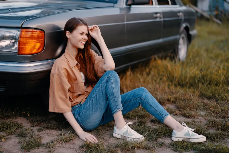 Young woman in a stylish outfit sitting near a vintage car, expressing joy in a natural outdoor setting. Summer vibes and casual fashion blend harmoniously.