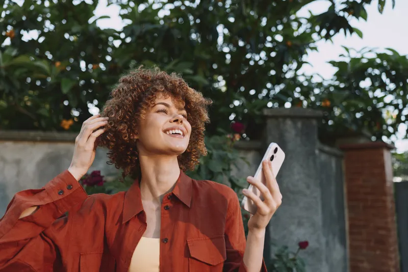 Young woman with curly hair smiles while taking a selfie outdoors, wearing a rust jacket over a light top, enjoying sunny weather and lush greenery around