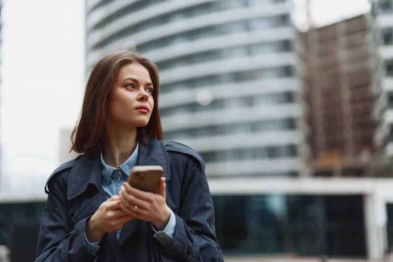 Thoughtful woman in a denim jacket stands outdoors in the city, holding a smartphone, looking away with calm, reflective mood and soft urban glow surrounds her.