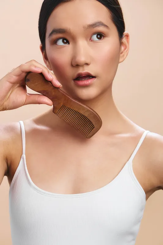 Asian woman stands in a neutral setting, holding a wooden comb to her hair, showcasing a calm grooming moment and personal care, with soft lighting and a clean aesthetic that emphasizes simplicity