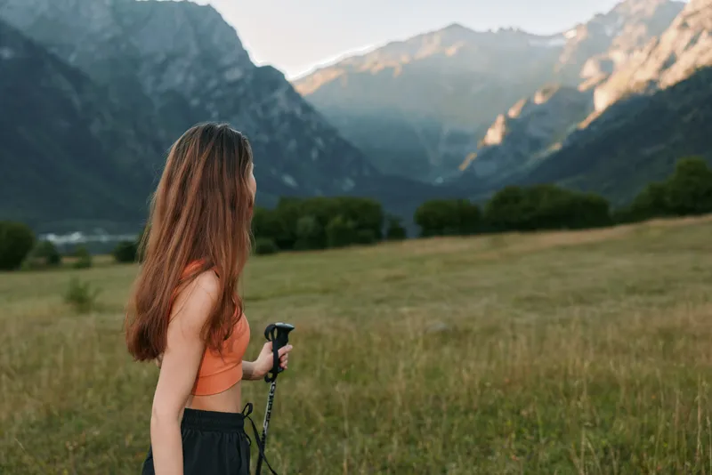 A woman stands in a grassy meadow facing distant mountains, holding trekking poles. She wears an orange top and dark pants, enjoying a serene outdoor landscape with fresh air and wide horizons.