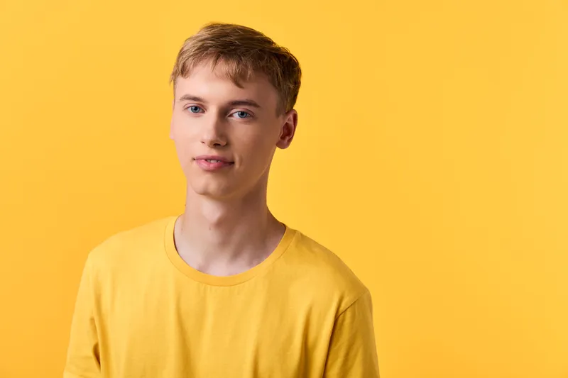 Young man wearing a yellow shirt stands in a studio against a bright yellow background, a cheerful smile showcasing casual fashion and modern lifestyle photography