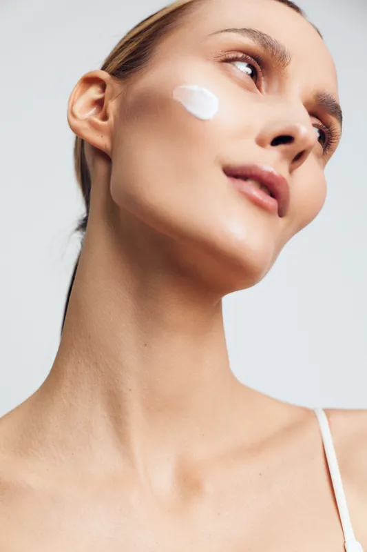 Young woman with clear skin applies white cream on cheek looks upwards with calm expression isolated on white background skincare beauty concept