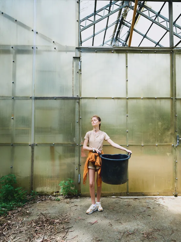 Woman in greenhouse holding a large black bucket, gardening and inspecting plants in casual outfit with jacket tied at waist, sunlight through glass panels and concrete floor.