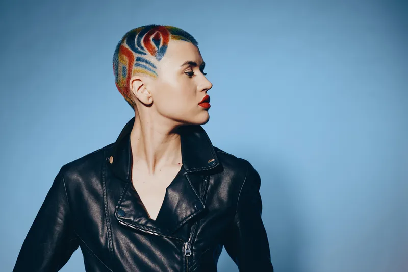 Woman with colorful shaved hairstyle and leather jacket posing against blue background, showcasing bold style and confident attitude in modern fashion portrait.