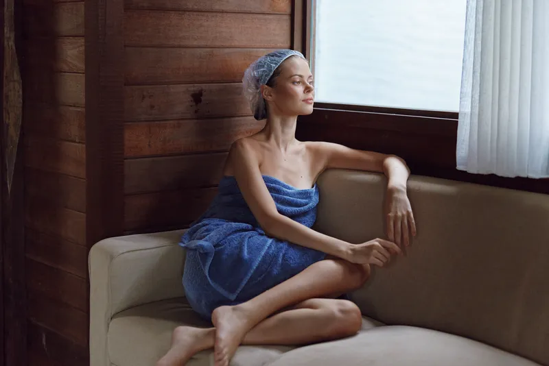 young woman sitting thoughtfully on beige sofa wrapped in blue towel with headscarf near window in wooden interior, natural light, calm atmosphere
