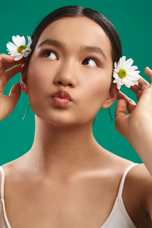 Asian woman with flowers in her hair smiles softly against a vibrant green backdrop, exuding freshness and natural beauty with a playful, artistic mood.