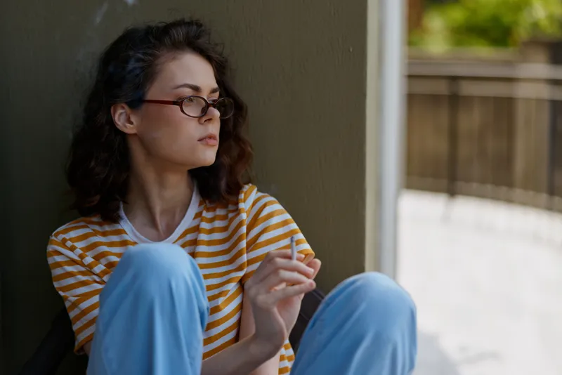 A thoughtful woman sits against a wall at home, wearing a striped orange and white shirt and blue pants, looking away with a calm, reflective expression.