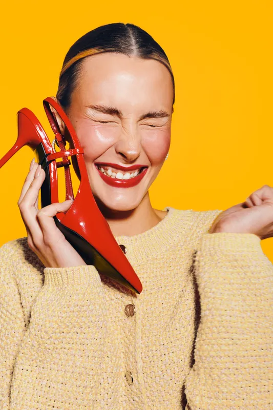 Happy woman with closed eyes holds red high heel shoes near face, smiling brightly on yellow background. Concept of fashion, joy, style, and vibrant emotions in studio.