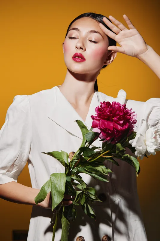Young woman with glossy red lips posing emotionally against bold orange background wearing white dress, holding vibrant red and white flowers. Concept of fashion, beauty, elegance.
