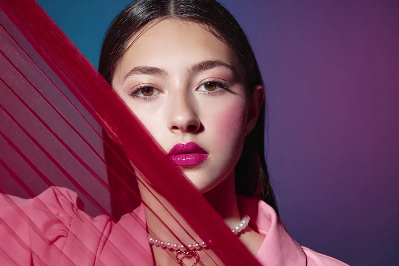 Young woman with vibrant pink lips and blush makeup poses behind a sheer red fabric. Close-up beauty shot on purple background. Concept for fashion, beauty, and creative styling.