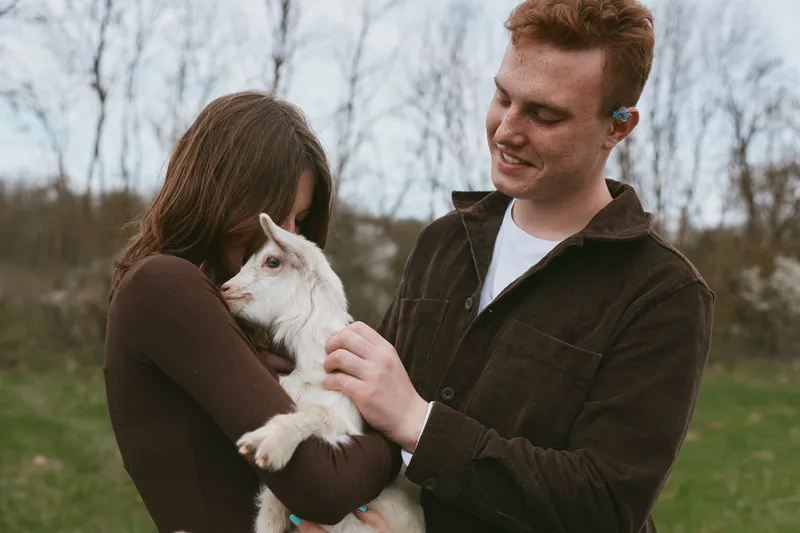 Young couple enjoys holding a baby goat outdoors in a natural setting during early spring, sharing a tender moment full of warmth and connection in casual clothing.