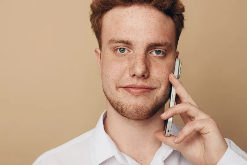 Young man with red hair and freckles talks on smartphone against beige background. Casual portrait of confident male using mobile phone for communication.