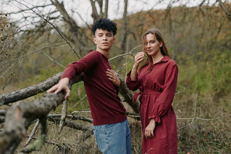 A young couple stands beside a fallen tree in an autumn field, a candid, genuine portrait capturing credible emotion and natural connection in an earthy outdoor scene with subtle fashion.