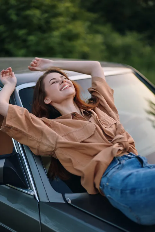 Young woman with long hair joyfully relaxing on a car, wearing a tan shirt and blue jeans, set against a lush green background, conveying happiness and freedom.