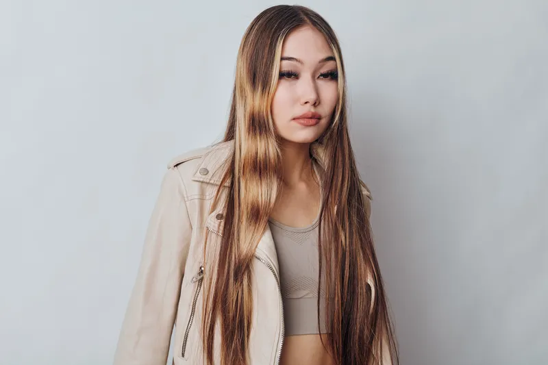 Young woman with long straight hair wearing light beige jacket and crop top posing against plain background in studio for fashion portrait photography.
