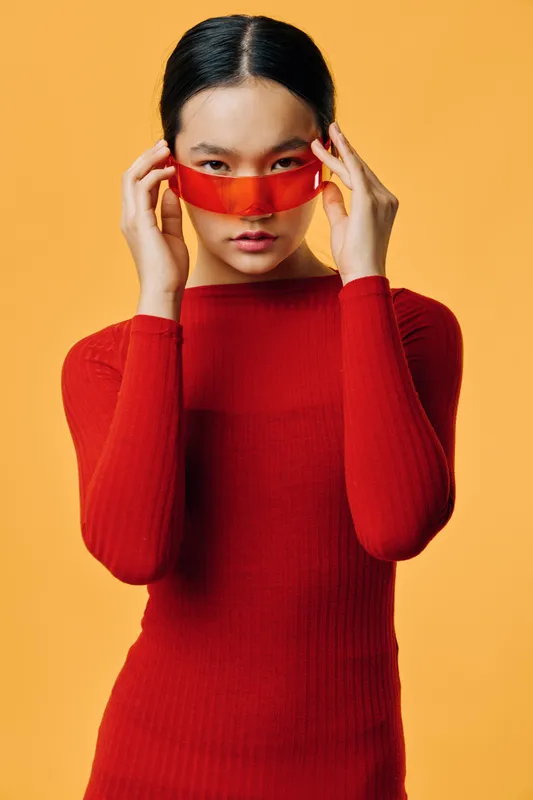 Young woman in red sweater adjusts futuristic red visor with hands on yellow background. Fashion portrait, modern style with intense gaze and confident expression in studio lighting.