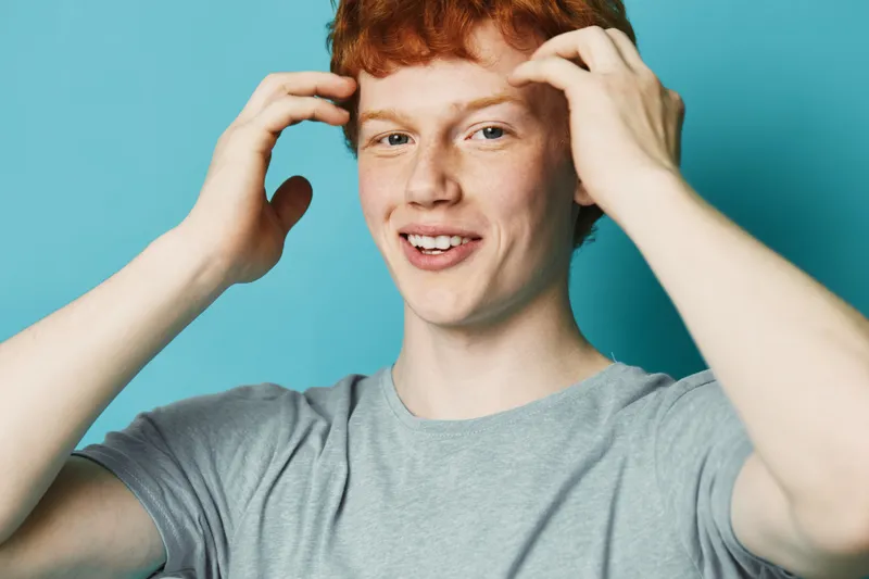 Young man with red hair and gray t-shirt smiling and touching his face on blue background. Portrait of happy male with freckled skin and casual style.