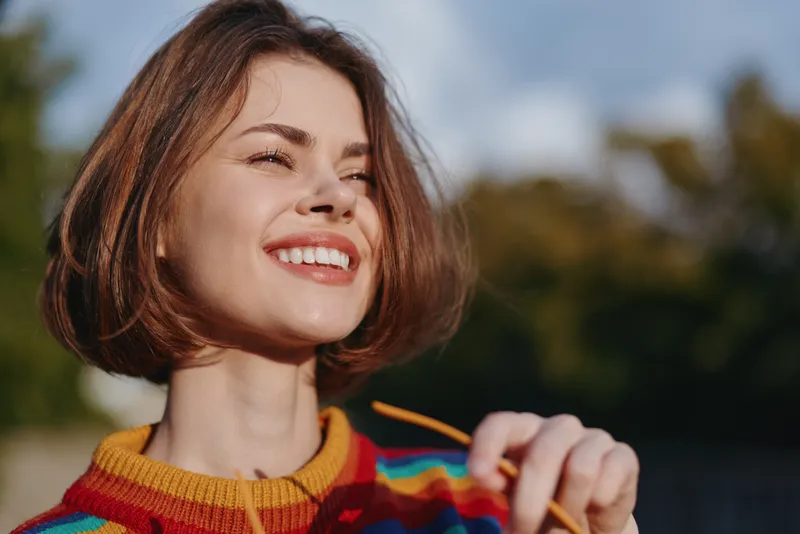 Smiling woman portrait outdoor colorful sweater sunglasses short hair, happy young woman enjoying sunlight in a closeup headshot, candid joyful expression and casual knitwear fashion vibe.