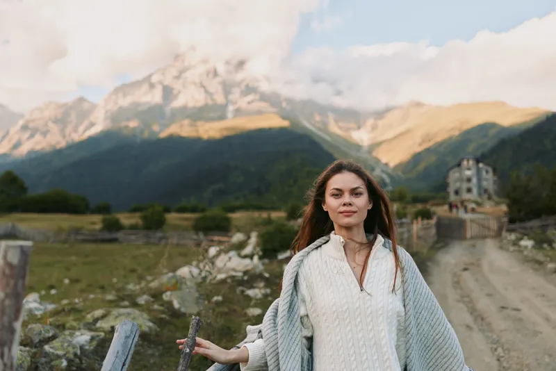 Woman with long hair wearing a white sweater stands before a grand mountainous landscape, with a stone wall and winding path behind her, conveying a cozy outdoor lifestyle and travel vibe.