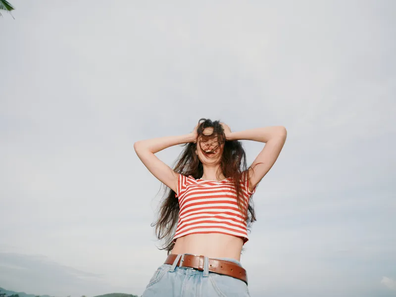 Joyful woman outdoors wears a striped crop top and jeans, arms raised, hair blown by wind as a bright sky frames a carefree, sunlit moment of youth and freedom.