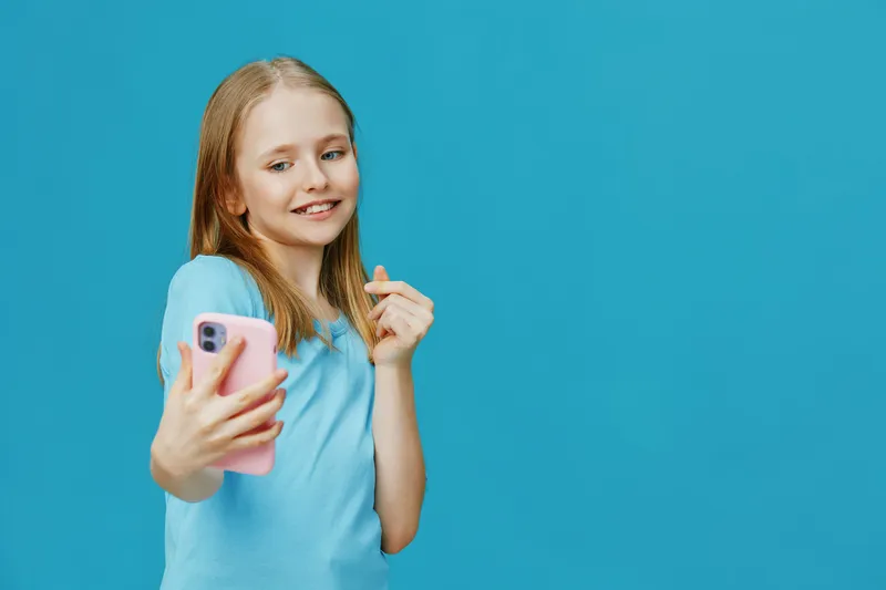 Young girl smiling while taking a selfie with a pink smartphone against a blue background, showcasing joy and playfulness in a bright and colorful setting