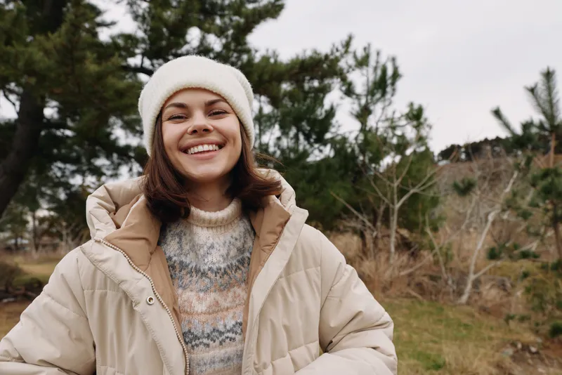 Woman in winter attire outdoors, smiling in a beige quilted jacket and white knit hat, standing among trees for a fresh portrait in chilly weather.